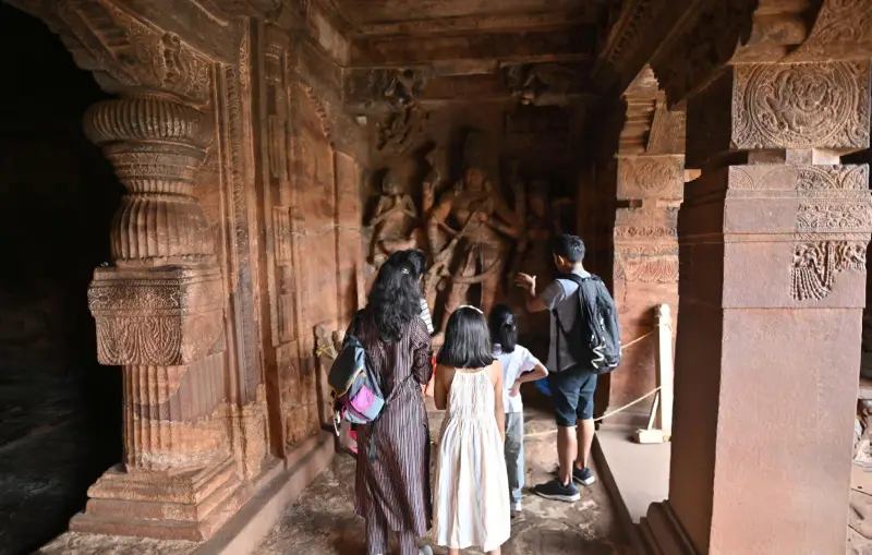 A tour guide explains an ancient stone carving to a family group at a historic Indian temple, showcasing intricate architectural details and cultural heritage.
