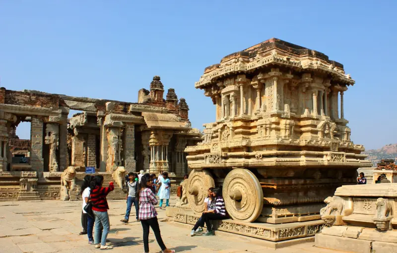 A group of tourists exploring the iconic Stone Chariot at the ancient ruins of Hampi, Karnataka, India, under a bright blue sky.